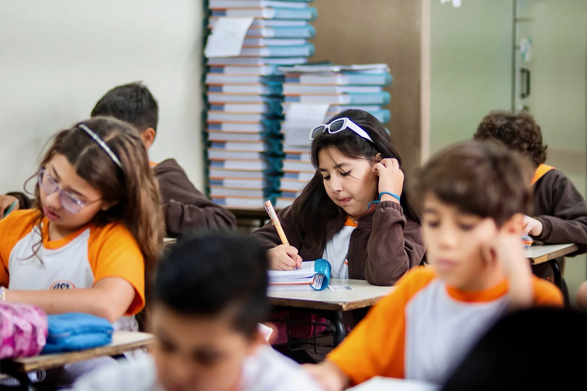 Alunos do Colégio OSE escrevendo no material didático, com livros empilhados ao fundo