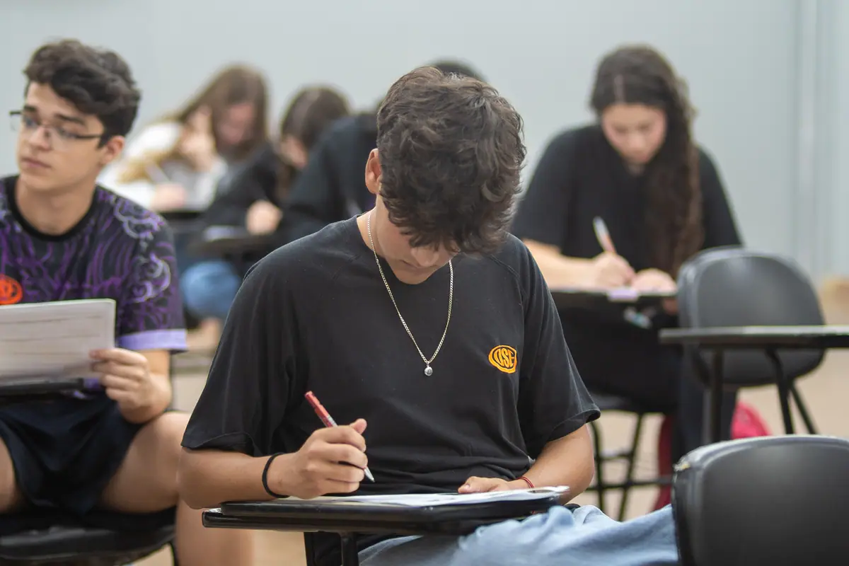 Aluno do Ensino Médio escreve na prancheta da carteira, ao lado dos colegas