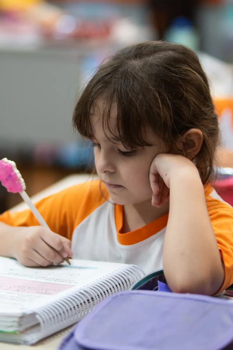 Menina pequena escreve no caderno espiral apoiando o rosto na mão