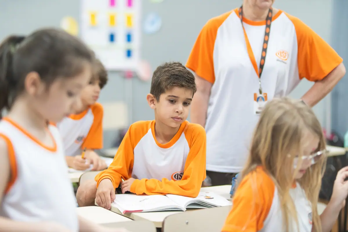 Professora acompanha em pé o aluno que estuda com o livro aberto na carteira