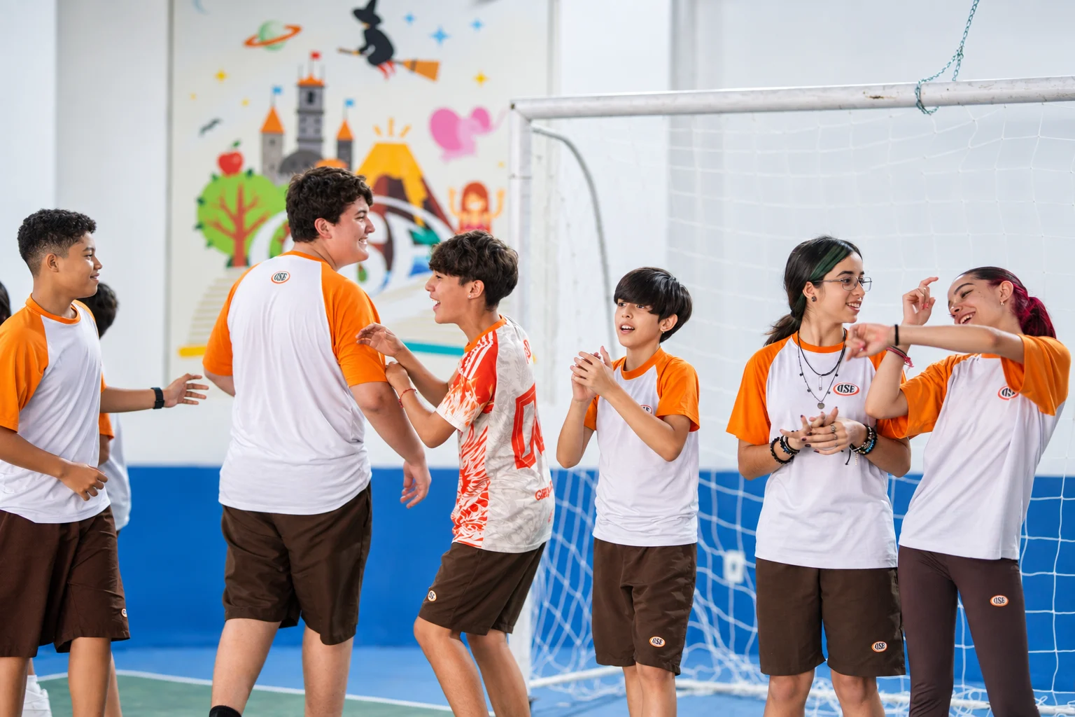Alunos riem e batem palmas em semicírculo numa dinâmica na quadra coberta