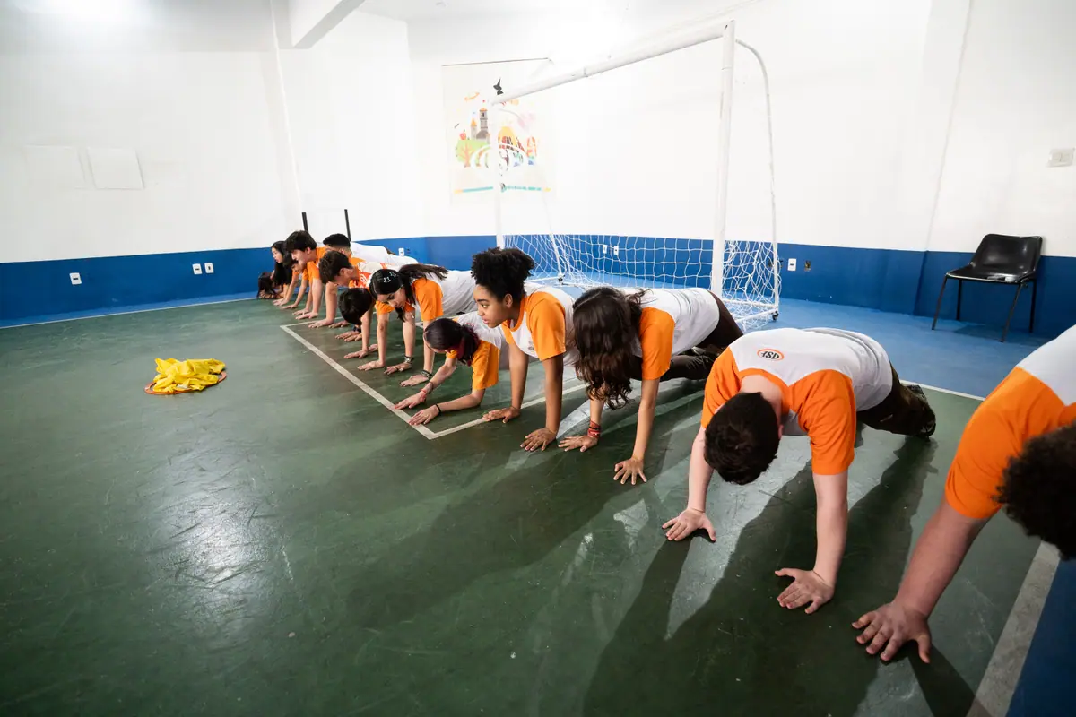 Fila de alunos em posição de prancha atravessa a quadra na diagonal