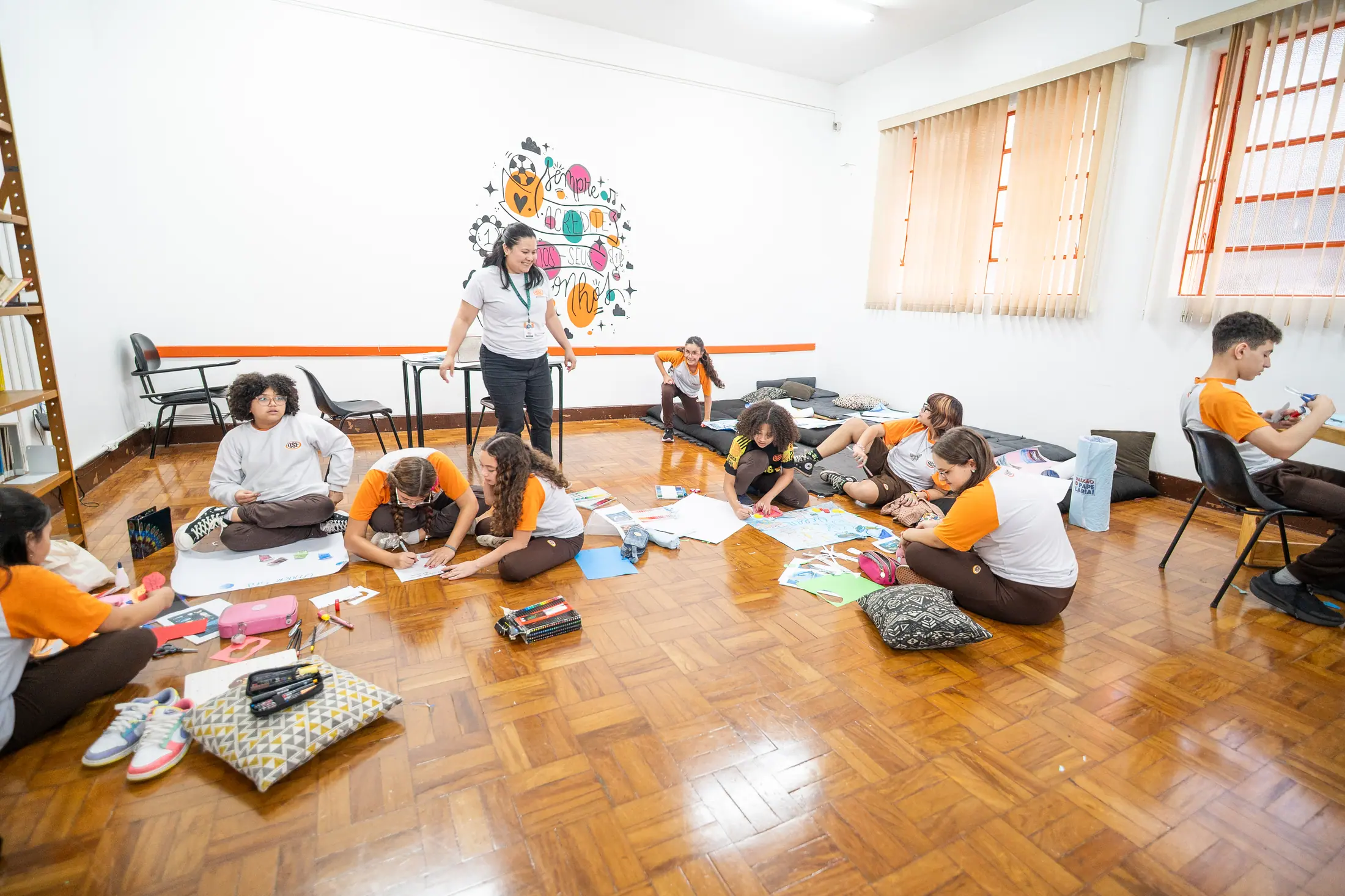 Alunos trabalham espalhados pelo chão da sala ampla, em pequenos grupos