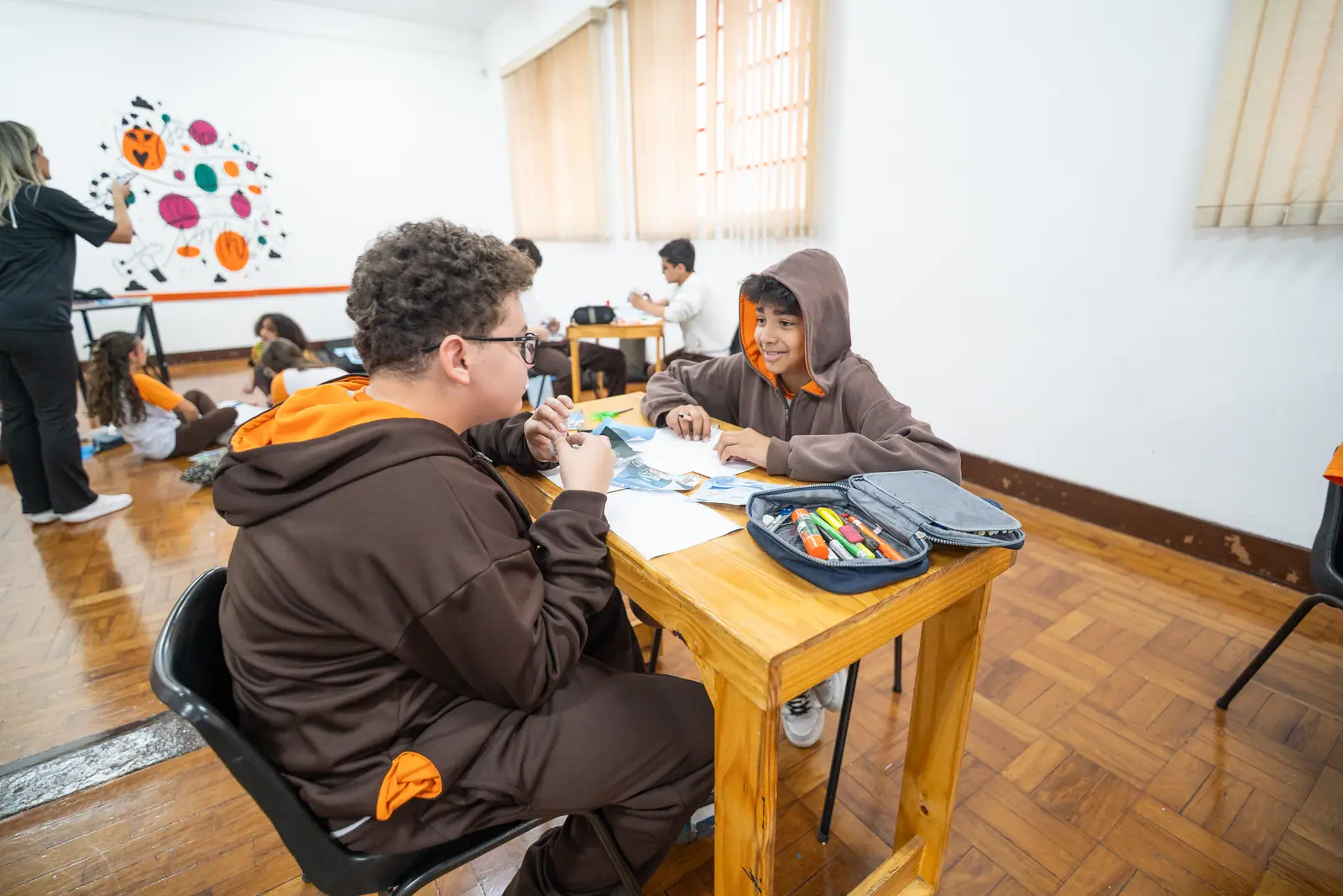 Dois alunos conversam e sorriem frente a frente enquanto recortam papéis