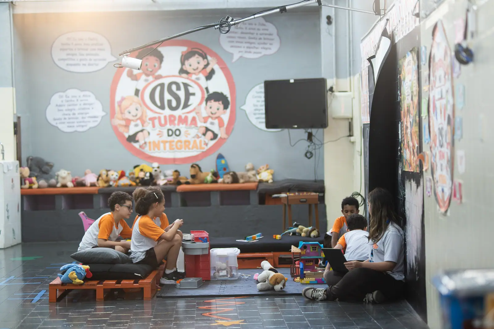 Crianças e monitora sentadas no chão da sala do Integral, sob o mural da turma