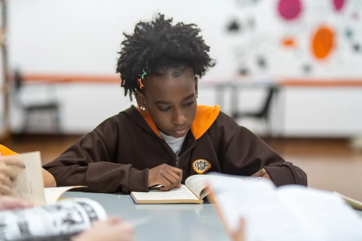 Aluna acompanha a linha do texto com o dedo e o lápis sobre o livro aberto