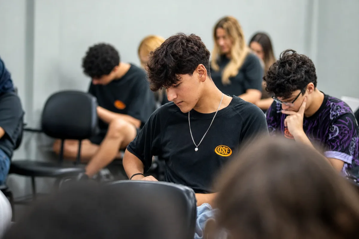 Jovem do Ensino Médio de camiseta com o brasão escreve na prancheta, no meio da turma